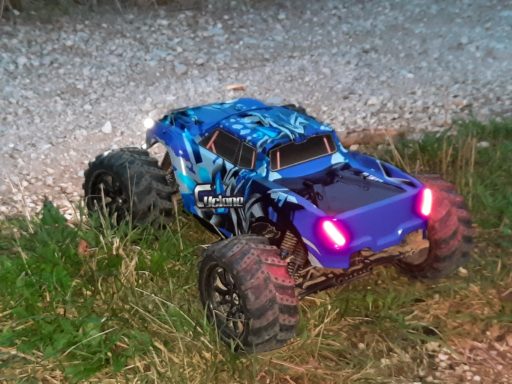Blauer RC-Monstertruck mit großen Reifen, steht im Gras auf einem Schotterweg.